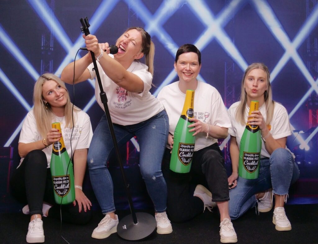 Vier fröhliche Mädchen bei einer Selfie Foto Party machen Spaß vor einer blauen Wand mit Mikrofon und Sektflaschen